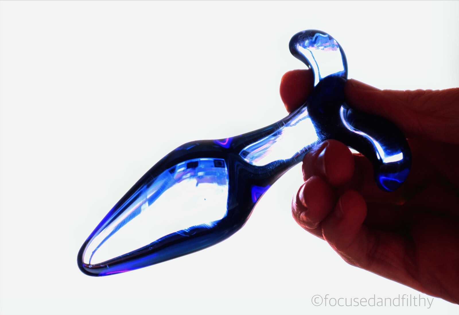 Colour photograph of a blue glass butt plug being held in a hand looking slightly see through