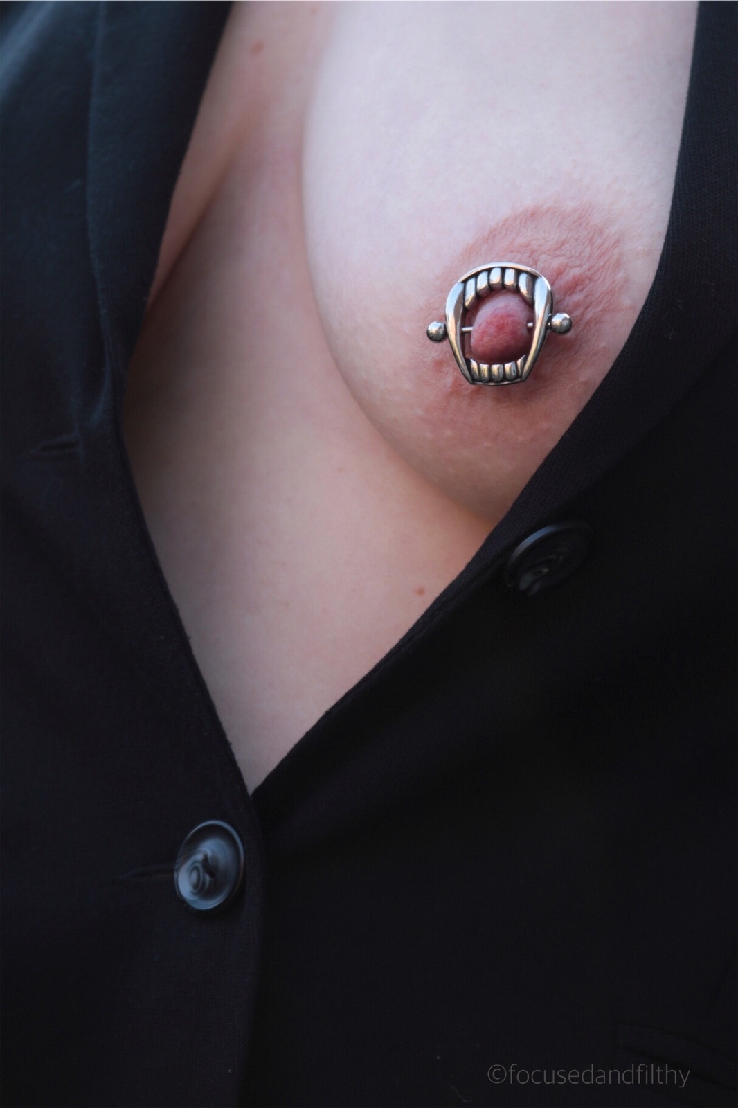 Colour photograph of a woman’s chest wearing a black jacket with her left naked breast exposed showing a pieces nipple with a large silver nipple bar with silver teeth around it. 