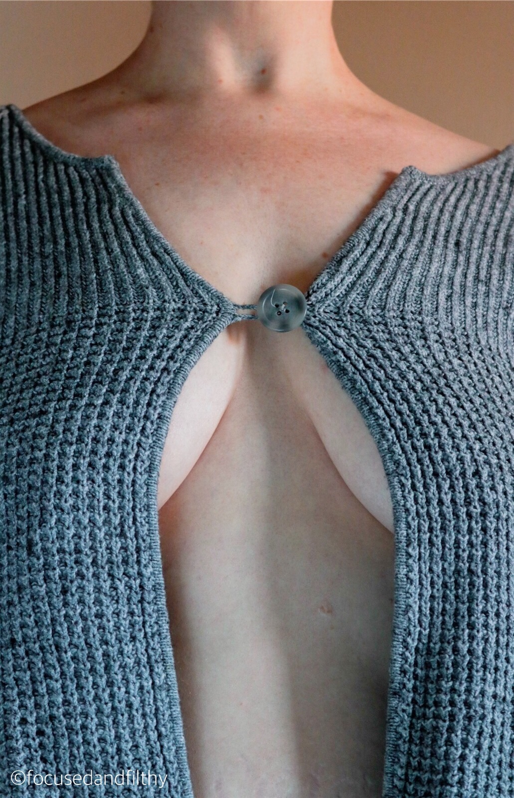 Colour photograph of a woman from shoulders to mid chest wearing j ust a grey cardigan held together over a cleavage with just one grey button