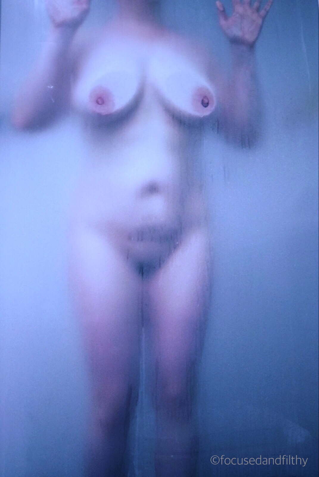 A colour photograph of a naked woman stood pressed up against a shower glass panel with a general blue colour to it 