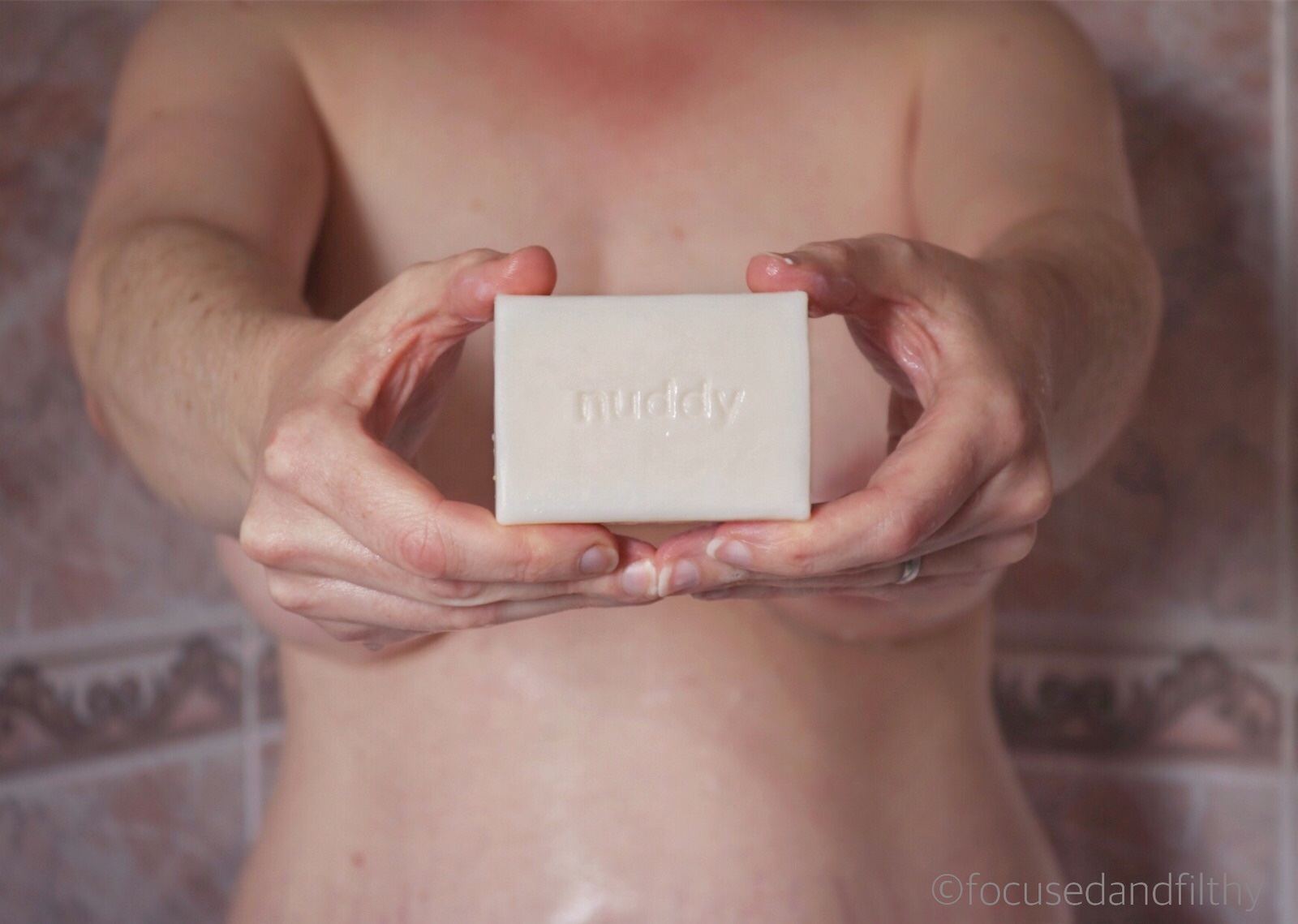 Colour photograph of a naked woman’s body in a bathroom and she is holding out a bar of soap closer to the camera which covers any of her breast. There is a word on the bar of soap which is Nuddy. 