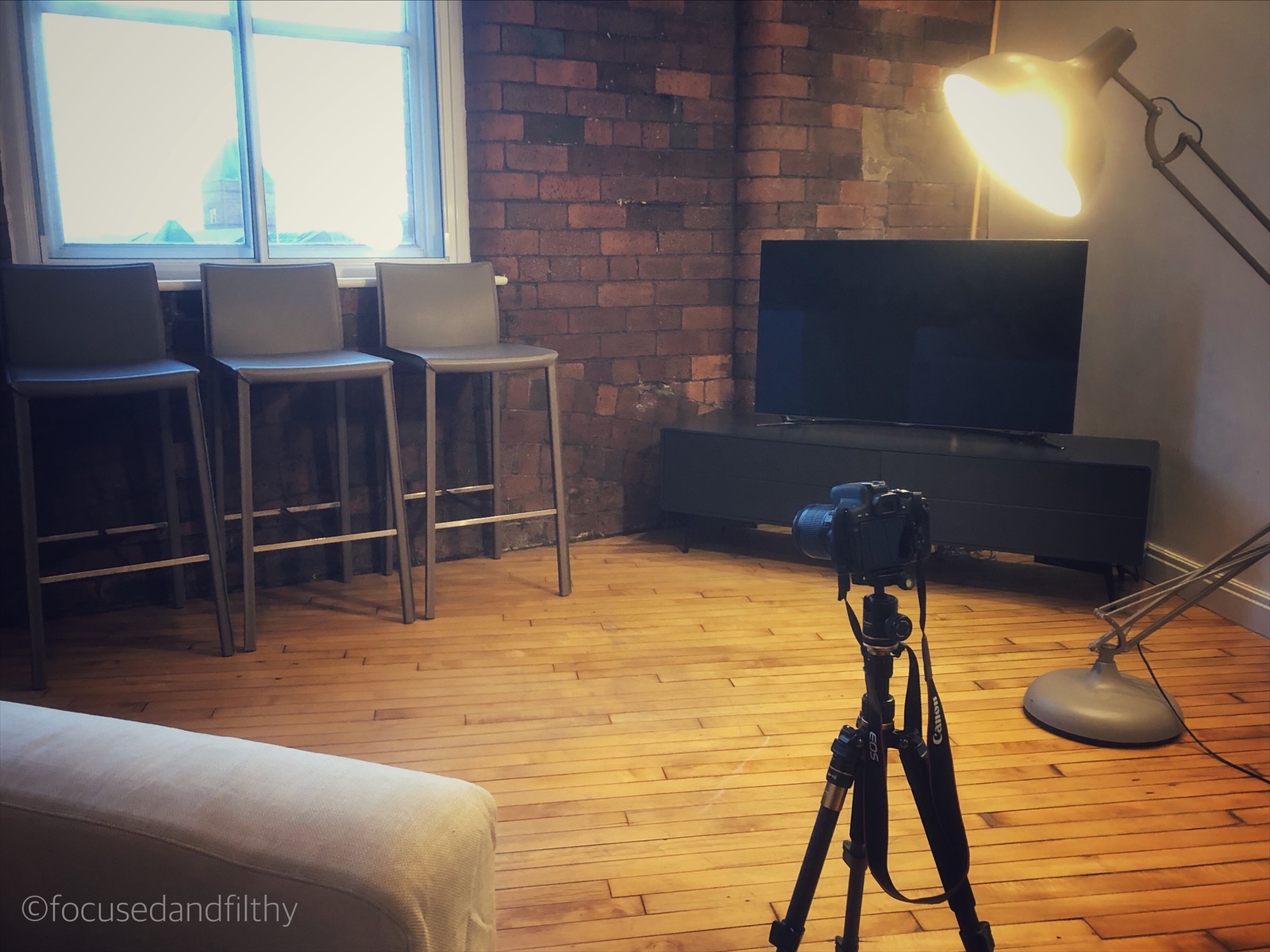 Colour photograph showing a camera on a tripod in a a large room with wooden floors and brick walls and three empty stools facing it 