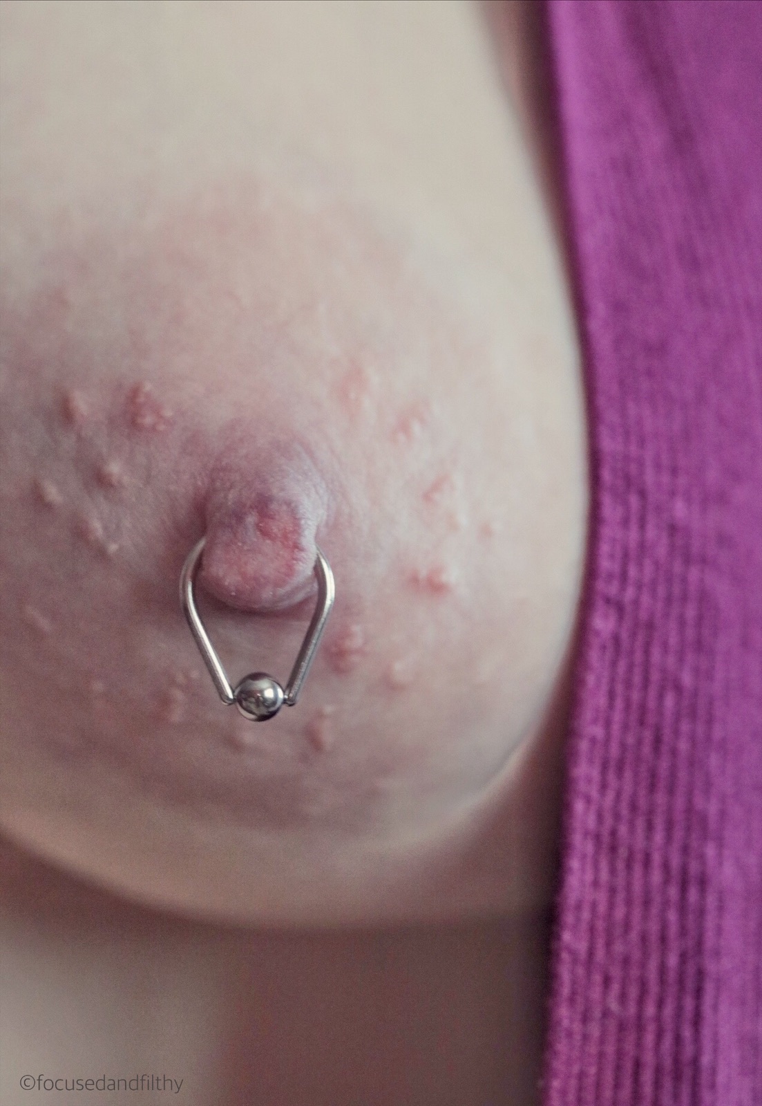 Close up colour photograph of a left nipple with a teardrop shaped nipple ring piercing. There is a bright pink cardigan seen to one side too  