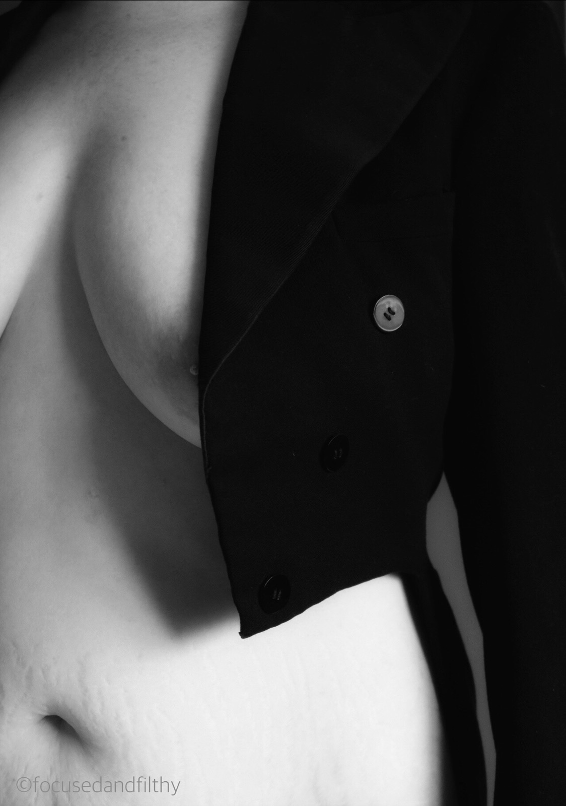 Black and white photograph of a woman from neck to waist showing her wearing just a tailcoat that just covers her left breast and shows the curve of her waist 