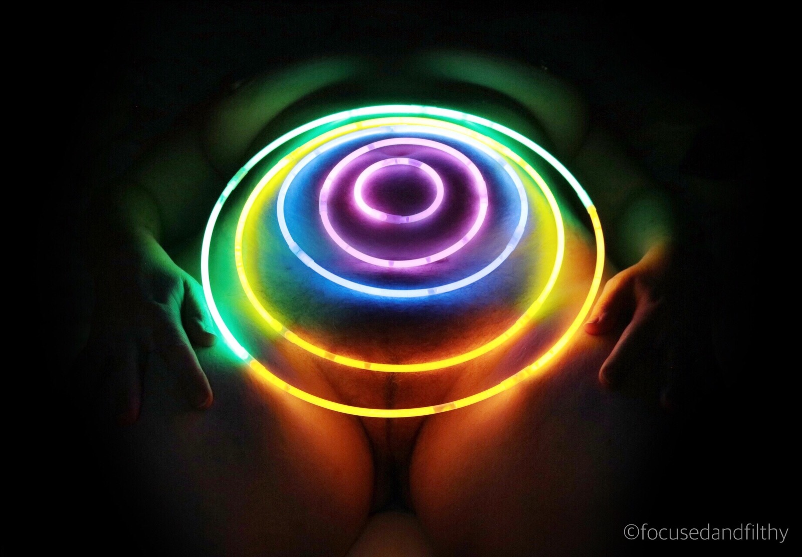 A rather dark Colour photograph of glow sticks made into different size circles and balanced on the stomach of a woman. The faint glow just lights up the edges of her body. 
