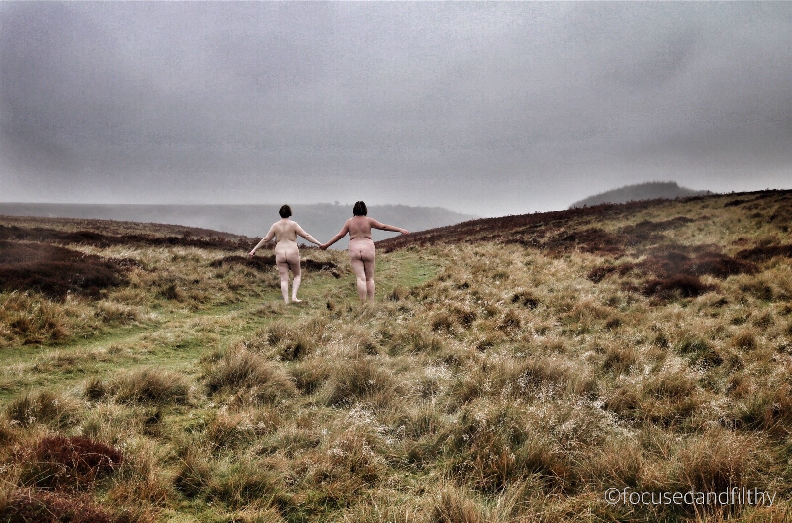 Colour photograph of the Yorkshire Moors on a grey wet day showing two naked women walking away along a grassy path with pale arses on show holding hands. 