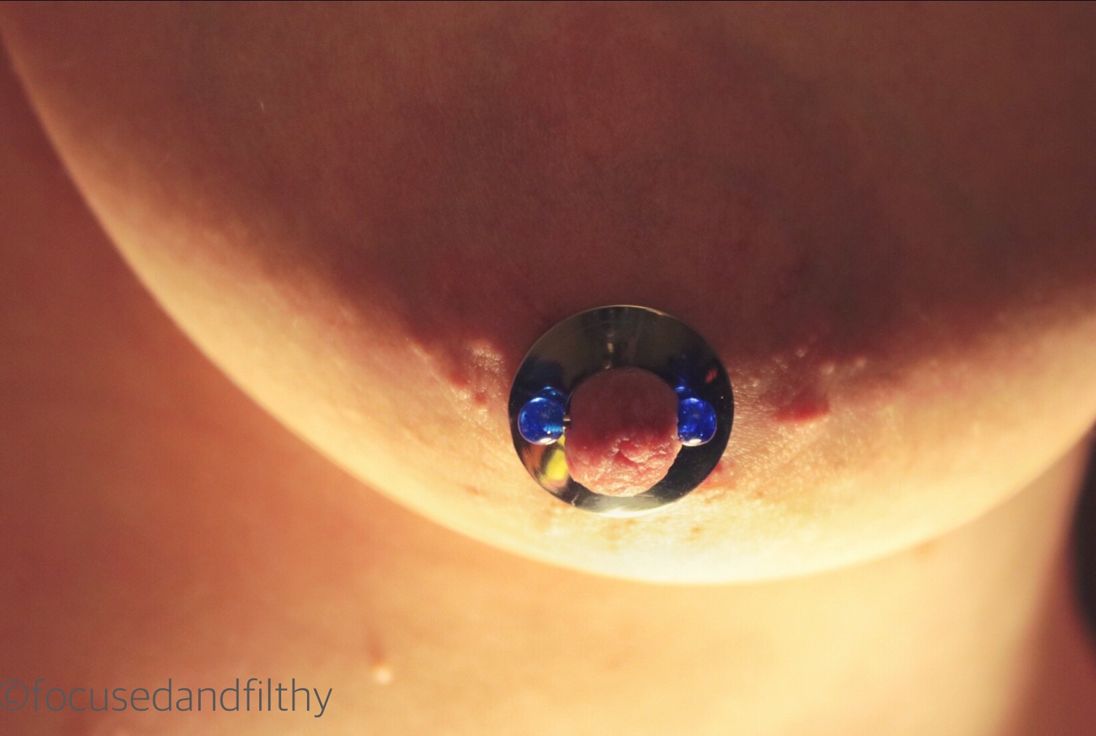 Close up colour photograph of my left breast showing a silver nipple shield held in place with a blue nipple bar lit from below 