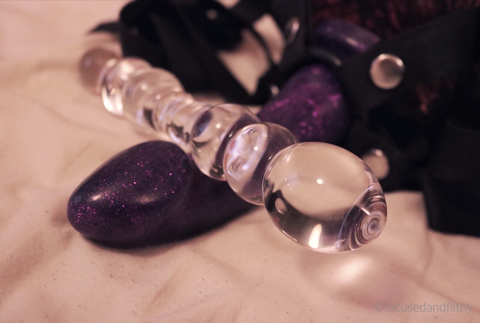 Close up colour photograph of a glass dildo laying across a purple sparkly silicone dildo in a strap-on laying on bedsheets 