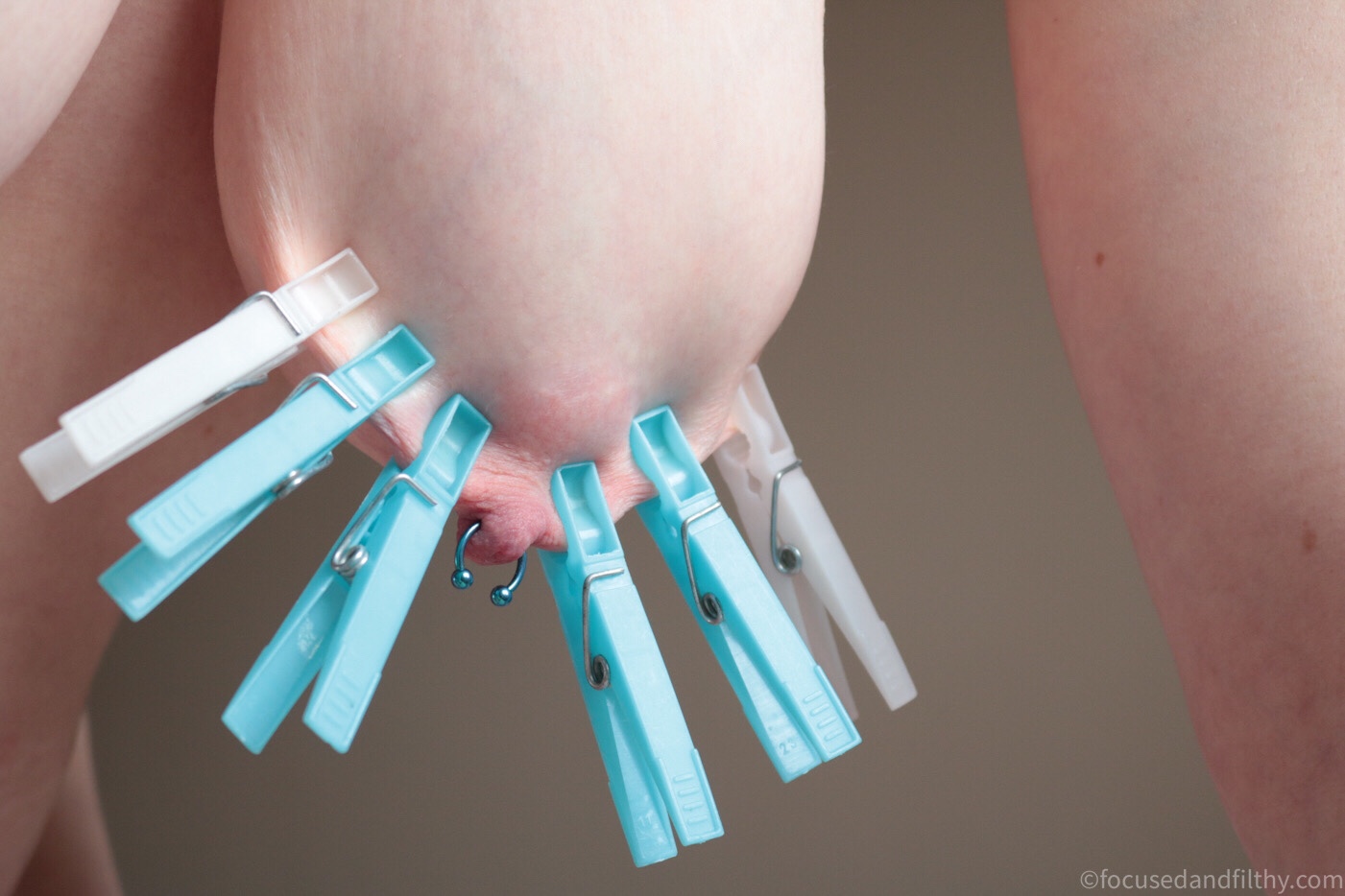 Colour photograph of a close up of a left breast hanging down (woman on all fours) the left breast has six blue pegs attached in a row hanging down in a fan shape 