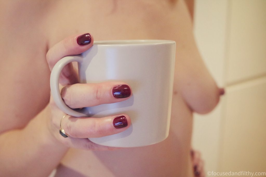 Close up colour photograph of a woman’s hand holding a mug of tea  while she is stood up obviously naked showing her bare breasts in a slightly blurred background  