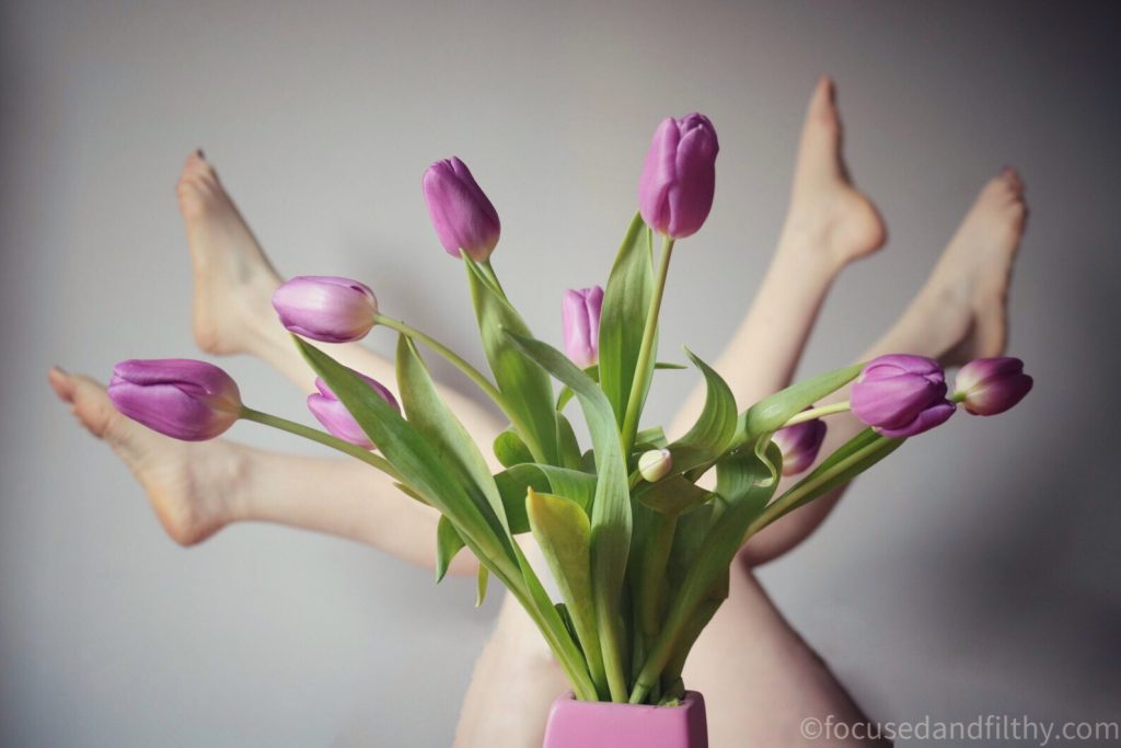 Colour photograph of a bunch of purple tulips in a pink vase and behind the vase are four bare legs sticking outwards mimicking the tulips stems and flowers  