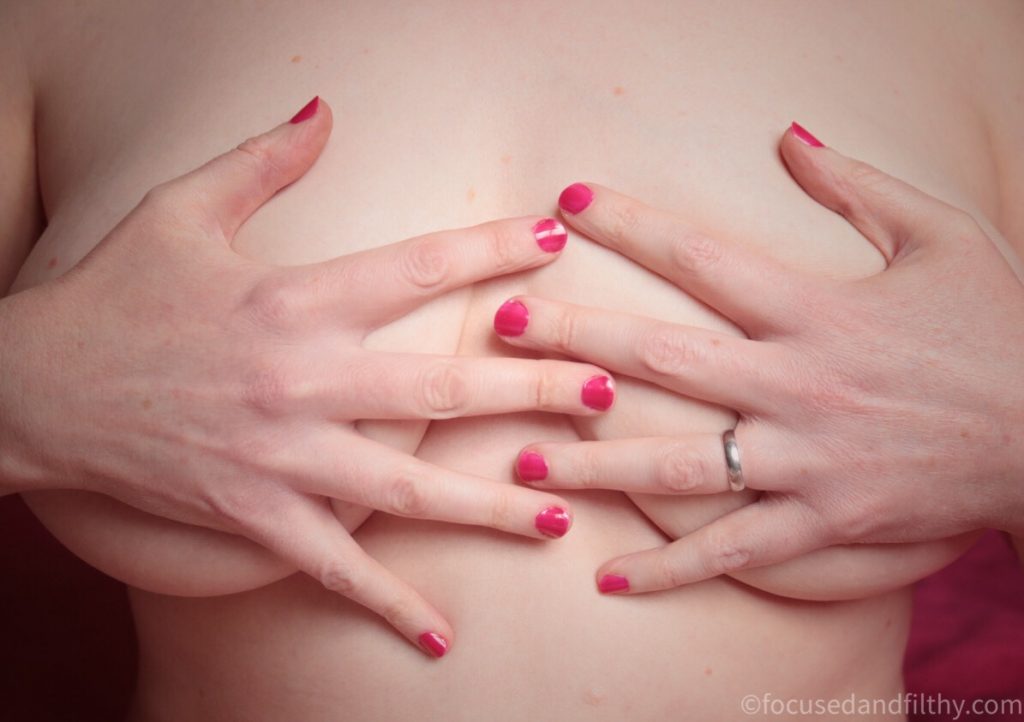 Close up colour photograph of two hands pressing down over two naked female breasts  the fingernails are painted a shocking bright pink  