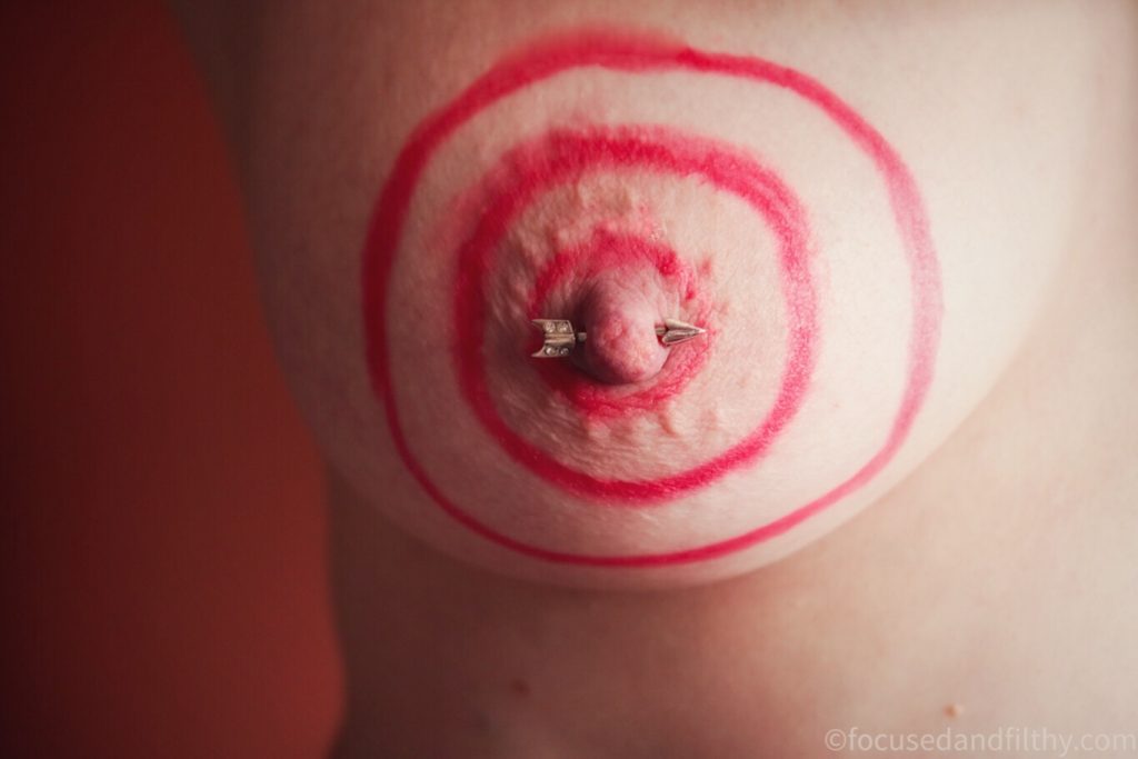 Close up colour photograph of a naked breast with red target rings draw round the nipple which is pierced with an arrow shaped nipple bar 