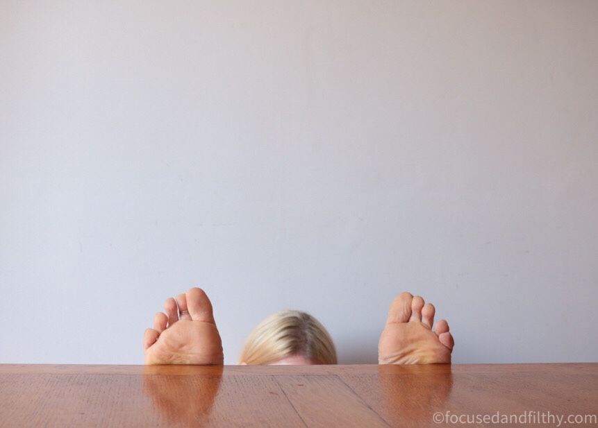 Colour photograph of a clear table top and just peaking over the top from below are two feet toes pointing upwards and the top of a head in the middle they must be sat on the floor below 