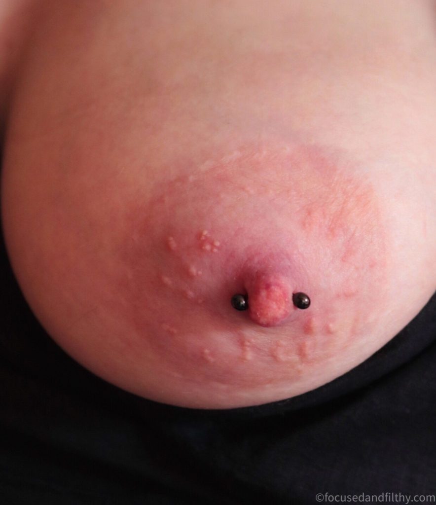 Close up colour photograph of a left naked breast with a small black nipple bar 