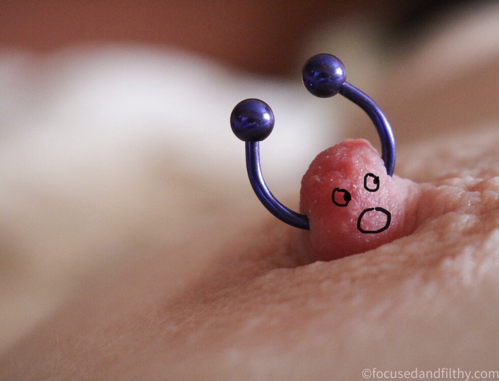 Close up colour photograph of a nipple with a horseshoe nipple ring in it standing upwards  it has been drawn on afterwards adding eyes and a mouth making the nipple ring look like arms thrown up in the air