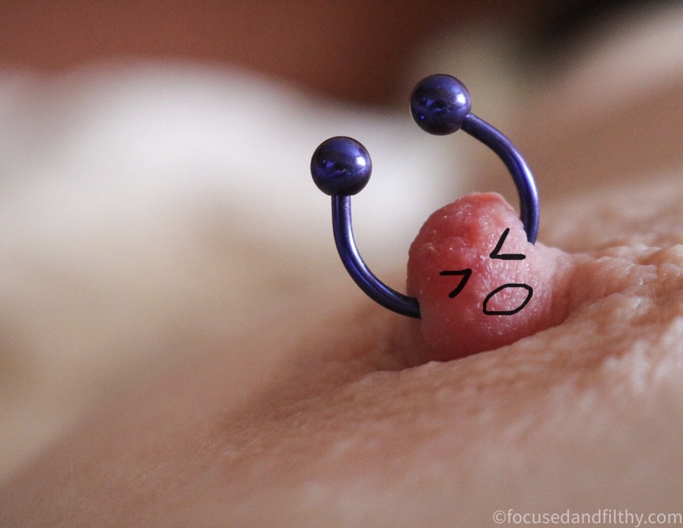Close up colour photograph of a nipple with a horseshoe nipple ring in standing upwards  it has been drawn on giving the nipple squinty shut eyes and a mouth so the nipple ring looks like arms thrown up in the air 
