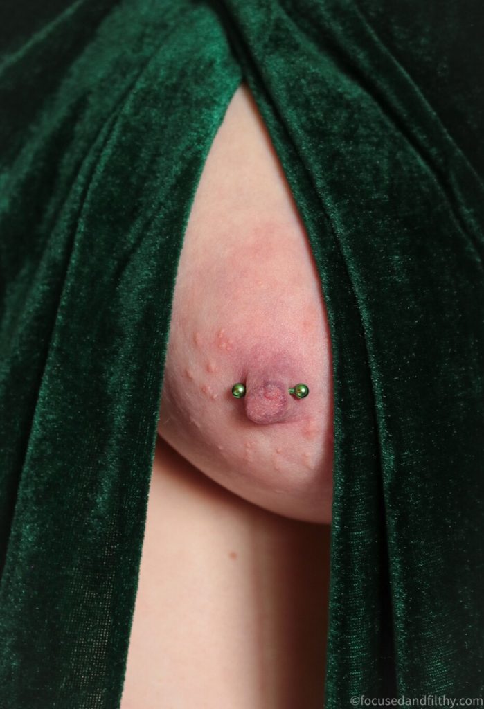 Close up colour photograph of a naked left breast draped with dark green velvet either side of it  the nipple has a green nipple bar through it 