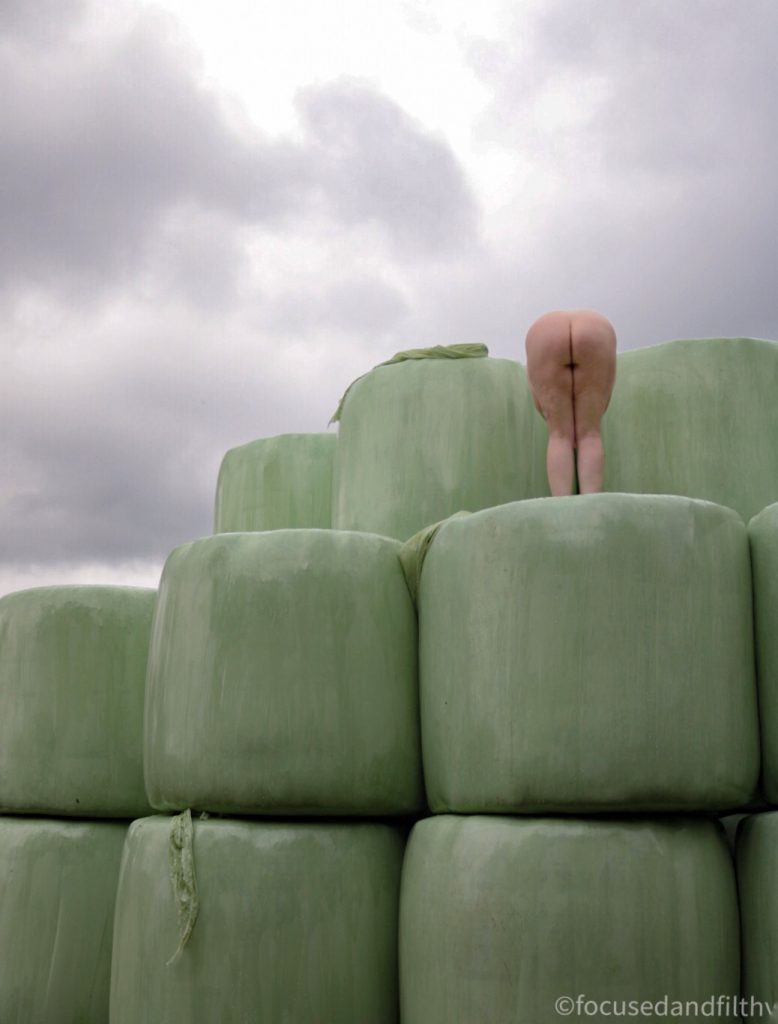 Colour photograph of some green plastic wrapped sillage bales with a naked woman stood on top  bending over showing her naked bottom 