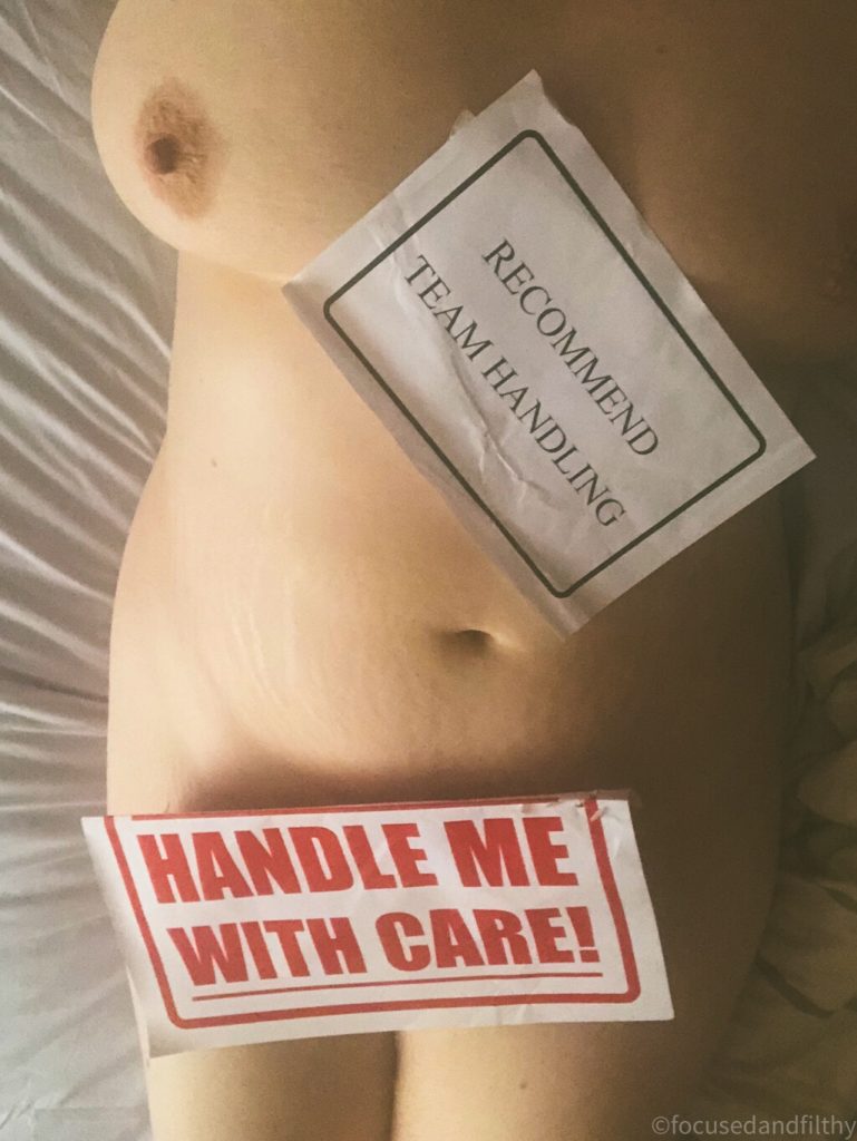 Colour photograph of the naked torso of a woman lying on a bed with two large stickers on  one saying handle with care, the other saying recommended team handling 