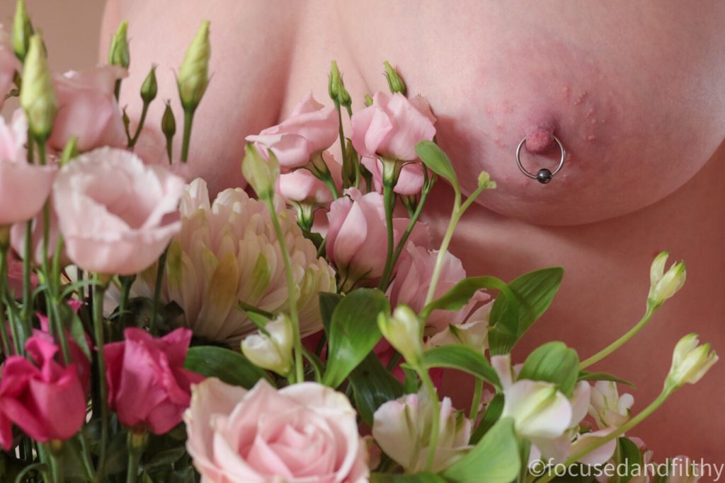 A colour photograph of a bouquet of pink flowers and behind is a naked female torso showing a left breast with a nipple pierced with a large silver ring with a hematite ball closing   
