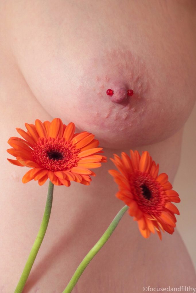 Colour close up photograph of a naked left breast with two orange Gerbera diasies held next to it and a red nipple bar though the nipple  