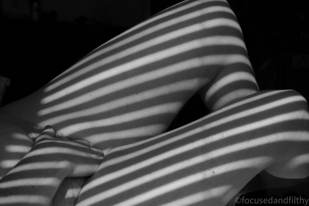 Black and white photograph of female legs in Venetian blind shadows and her hand between her legs   