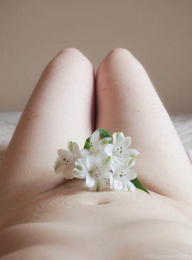 Colour photograph which is from the photographers perspective looking down a naked body at some white flowers covering her muff and her pale knees up in the air   