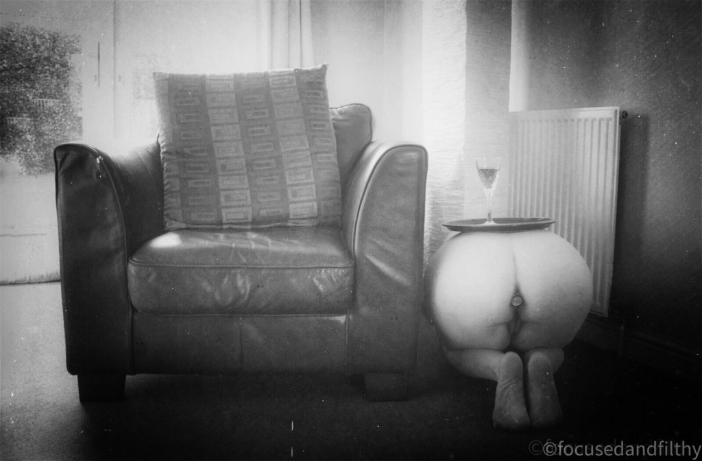 Black and white photograph of a single sofa seat and next to it is a naked woman with a tray on top and a wine glass acting as a side table  only her bottom is on display 