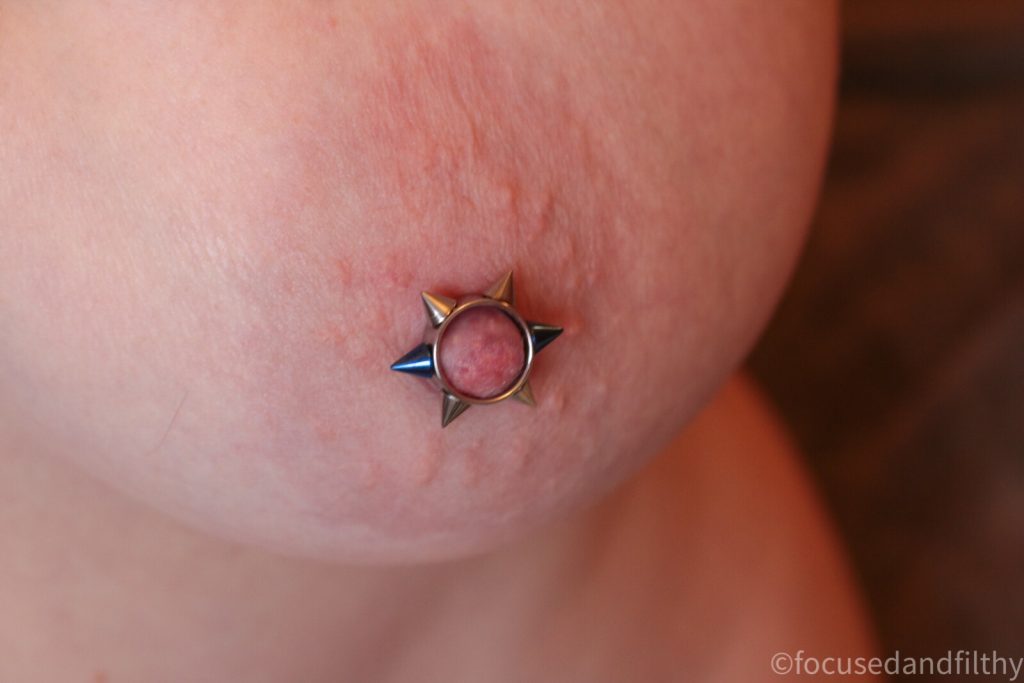 Close up colour photograph of a left naked breast with a nipple cuff with 6 metal spikes round it  two of which are blue 