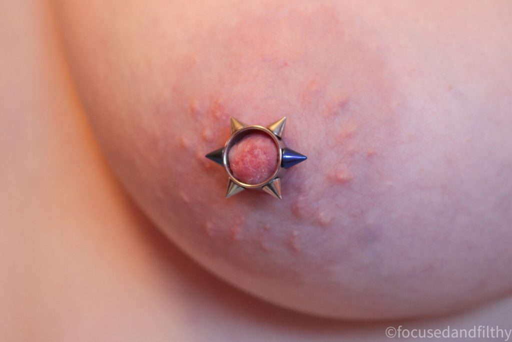 Close up colour photograph of a left naked breast with a nipple cuff with 6 metal spikes round it  two of which are blue 