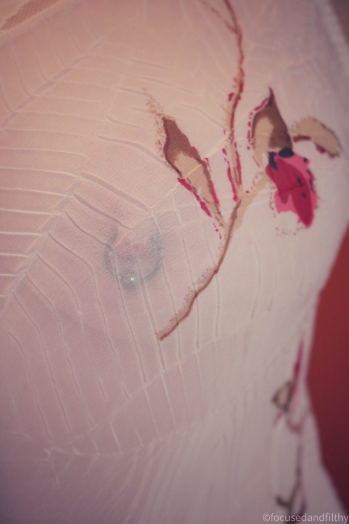 Colour photograph of a sheer white camisole and underneath you can see a large black nipple ring in the nipple 