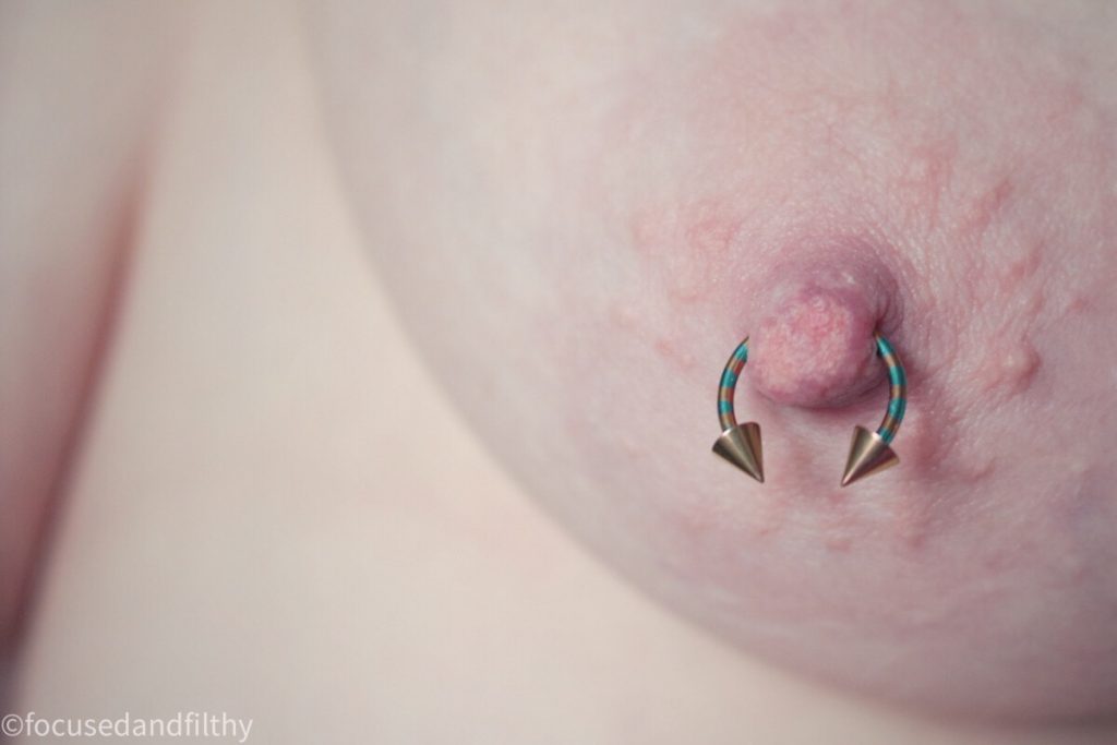 Close up colour photograph of a pierced nipple with a horseshoe ring that is striped in turquoise and bronze with cone points on the ends 