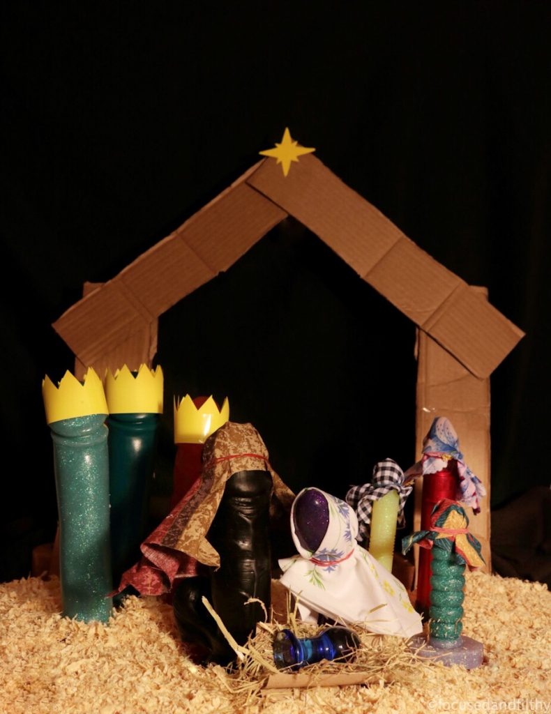 Colour photograph of a dildo nativity scene including three tall dildos wearing crowns, three smaller disposable wearing tea towel like head dressers as shepherds, Joseph is a large black dildo with balls and Mary is a purple sparkly dildo with a white shawl , Jesus in a manger is a small blue glass butt plug it is staged on a dark night in a cardboard stable with a bright star above and sawdust all around 