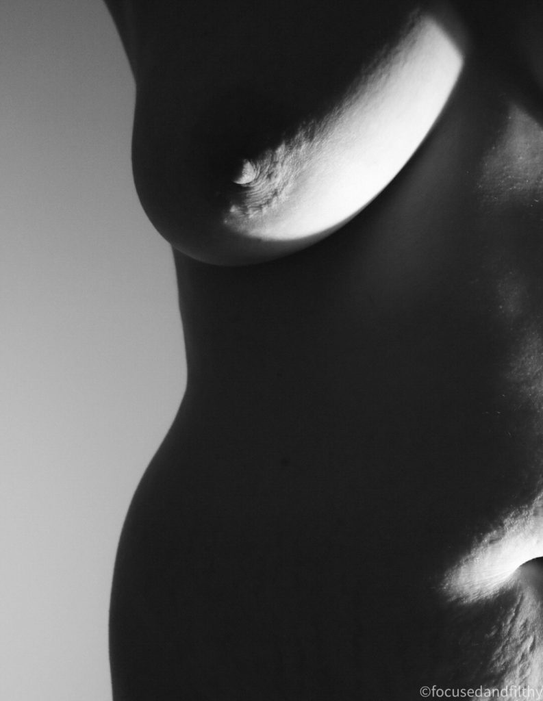 Black and white photograph of half a naked woman torso only lit from below showing a dark curve of the hip and only light catching her right breast  