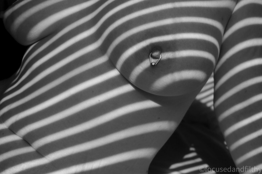 Black and white photograph of a naked woman’s chest with the shadowy stripes of a blind running diagonally across her 