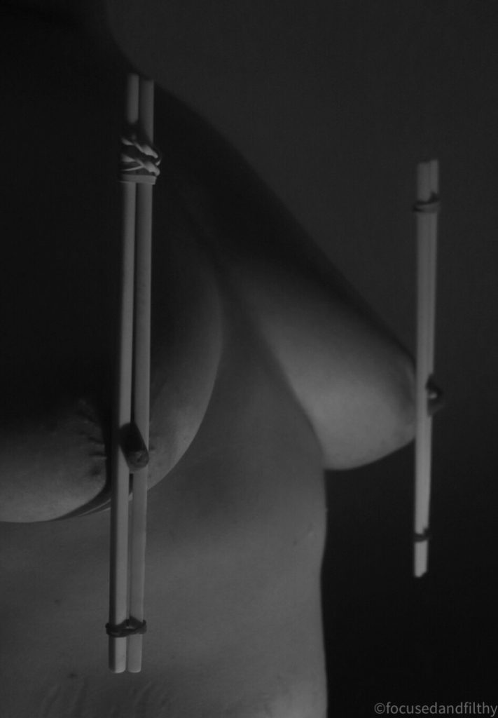 Black and white photograph of a naked woman’s chest showing pairs of chopsticks vertically held together with elastic bands at the end clamping both nipples in the middle 