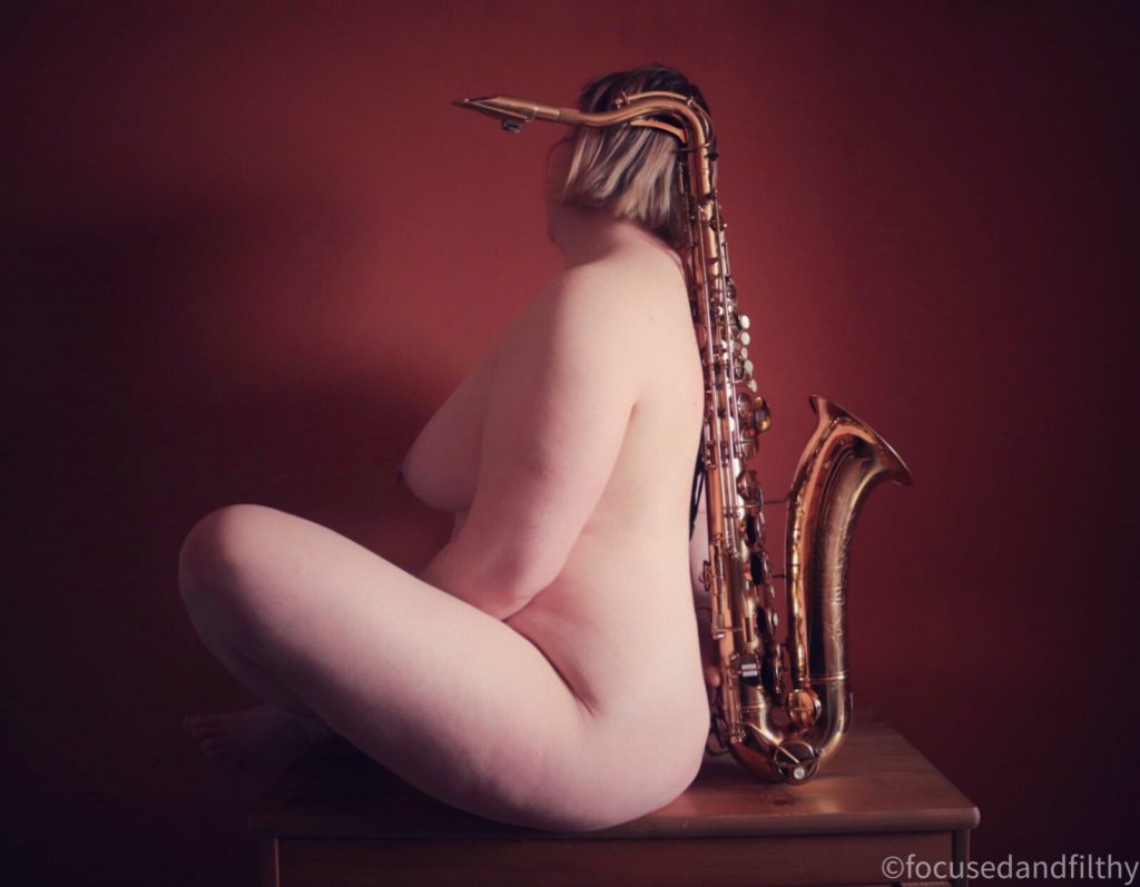 Colour photograph of a naked woman sitting with her back to a tenor saxophone  her shaped reflecting that of the instrument 