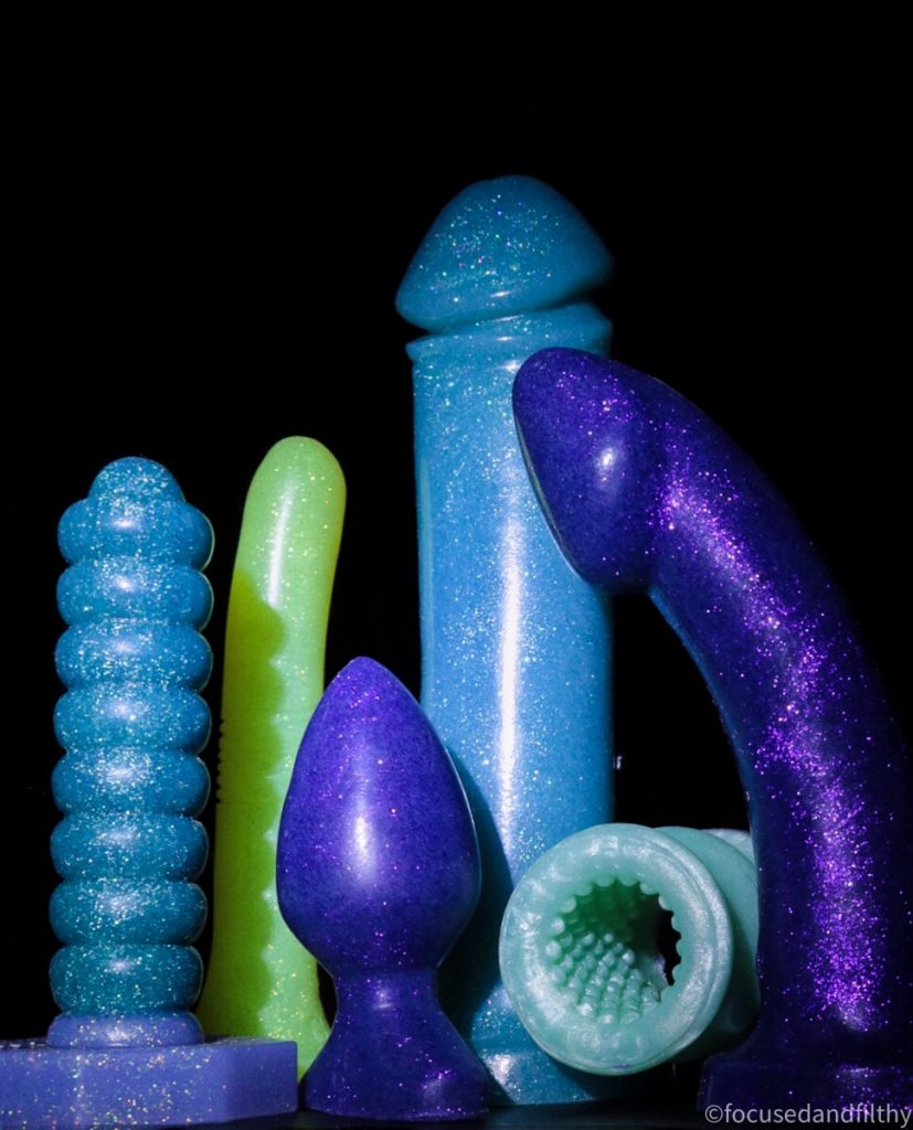 Colour photograph of several sparkly different shapes dildos and butt plug with a dark background  the toys are mainly turquoise and purple 