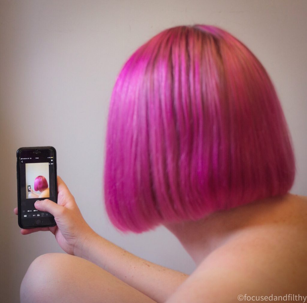Colour photograph of the back of a pink Bob hair and holding a mobile phone that shows the same image of the back of the pink hair 