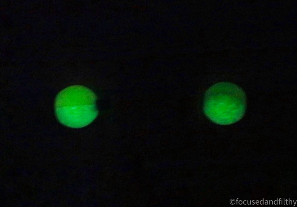 A photograph of a black background with two spheres of green shown in a horizontal line in the middle (which are the two glow in the dark nipple balls on a nipple bar)