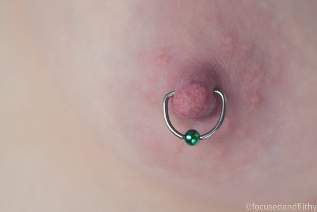 Close up colour photograph of a left nipple with a silver D shaped Nipple ring in with a bright green ball closure on it  