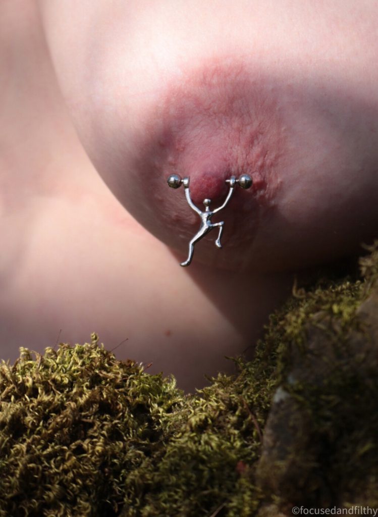 Colour close up photograph of a naked left breast above a missy drystone wall with a nipple bar in and a small silver man dangling from it looking like he’s climbing up 