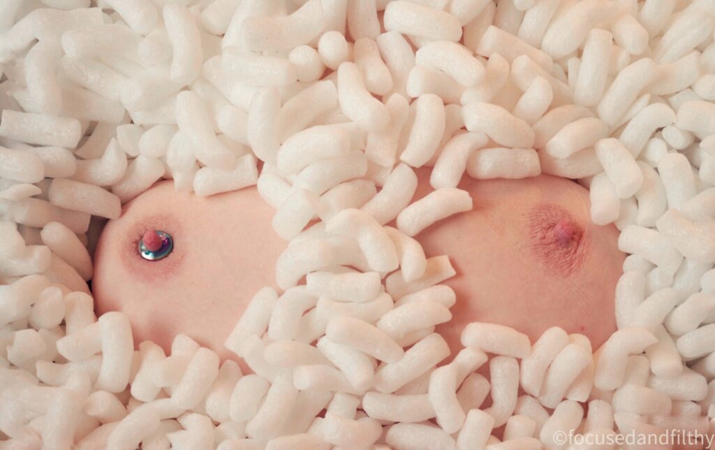 A colour photograph of a load of white packing foam beans with two pink boobs sticking up in them (but you can’t see any other part of a person) 