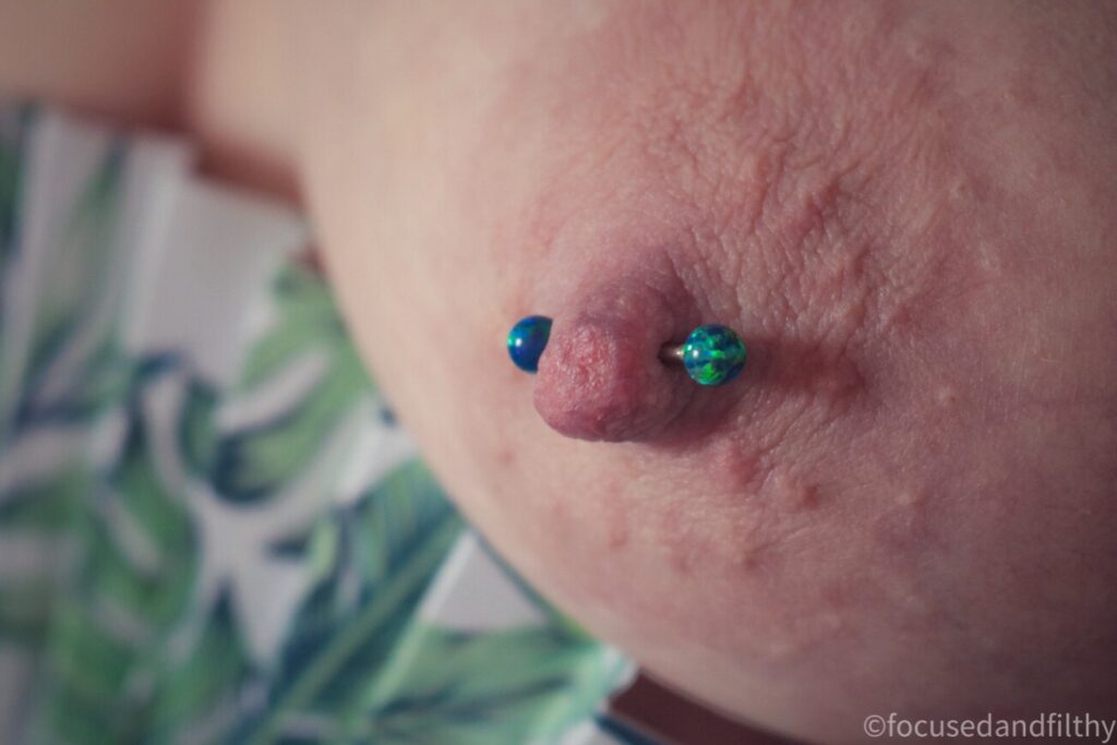 Close up colour photograph of a naked left breast with a green abs blue Opal style balls on a nipple bar and covering the right breast is a white paper and wood fan with a green leaf pattern on 
