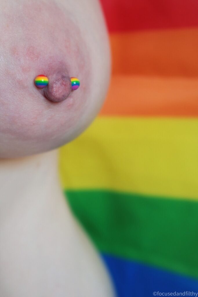 Close up colour photograph of a left naked breast with a nipple bar in which has rainbow stripes bows on each side. The background is the pride rainbow flag too 