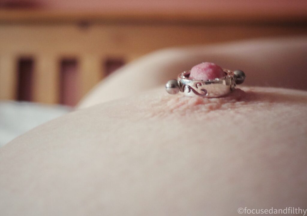 Close up colour photograph of a naked left breast looking down at the nipple with a silver decorated nipple cuff on 