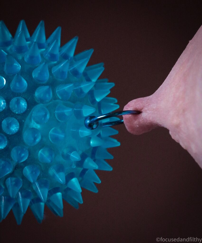 Close up colour photograph of a nipple being pulled by its blue nipple ring that is attached to a blue spiky ball