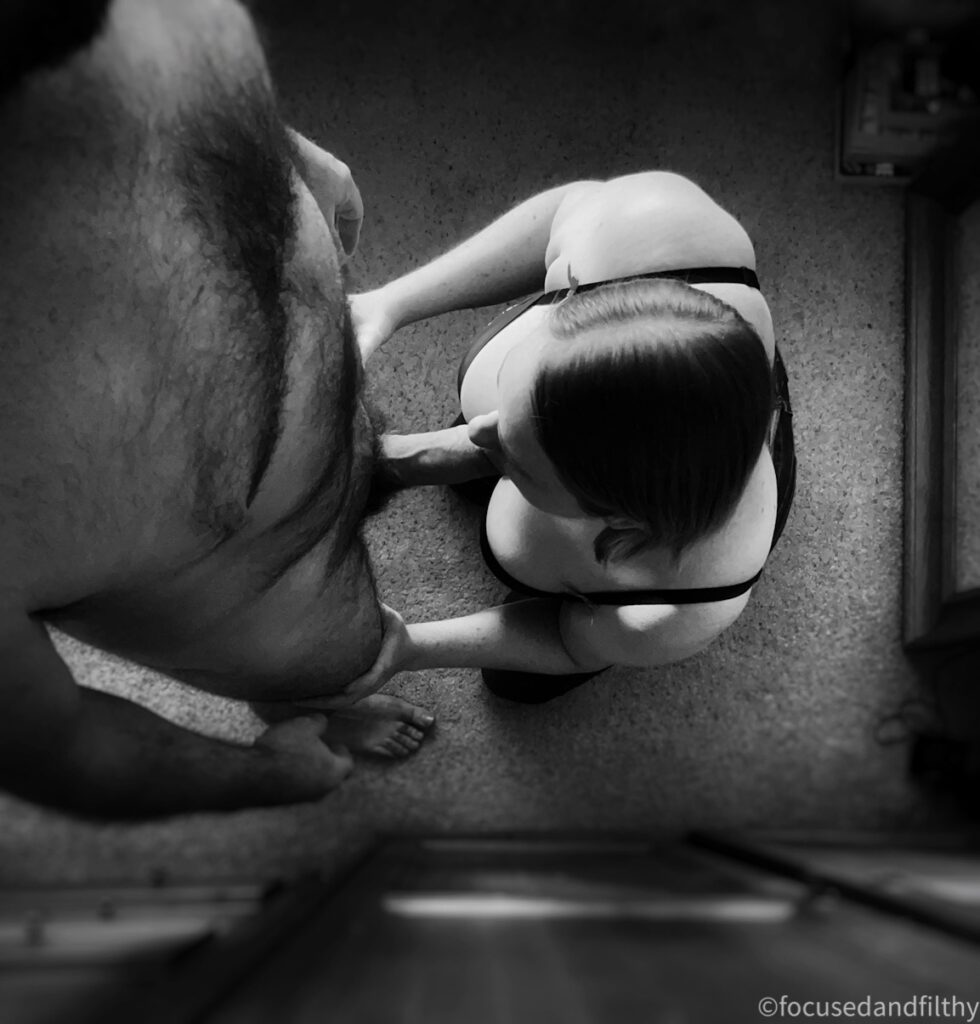 Black and white photograph taken from above showing the head of a woman on her knees sucking on the cock of a man stood next to her with a mirror behind her  