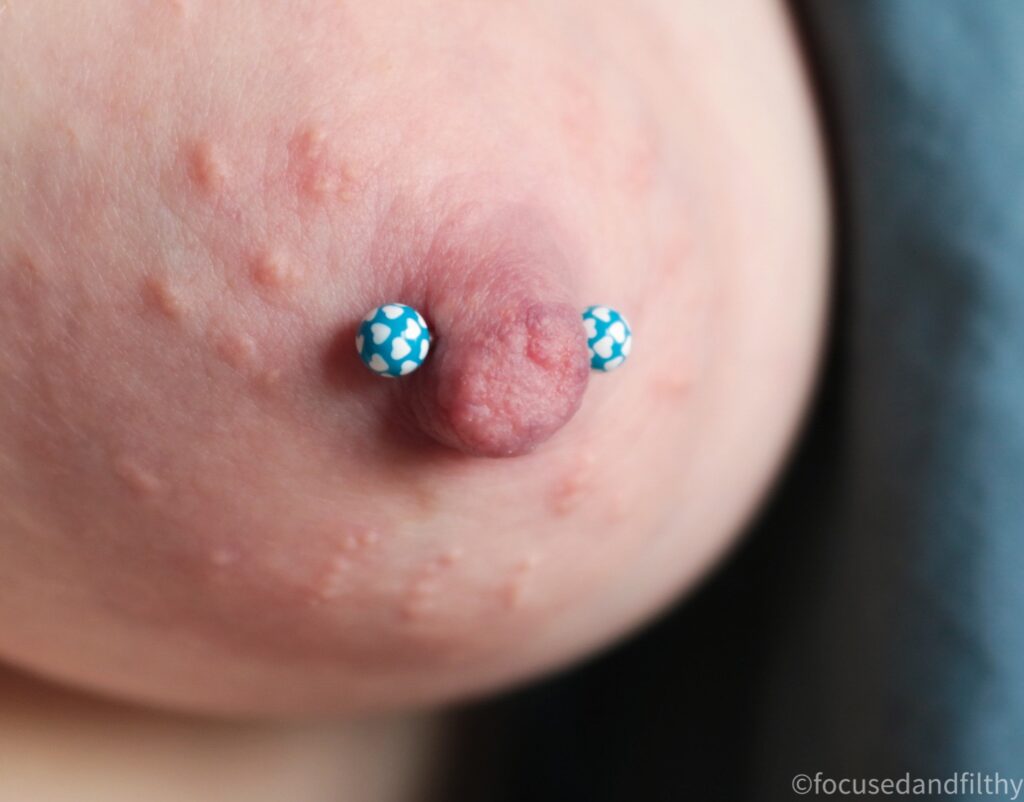 Close up colour photograph showing a naked left breast with a blue nipple bar that has little white hearts all over the blue balls. 